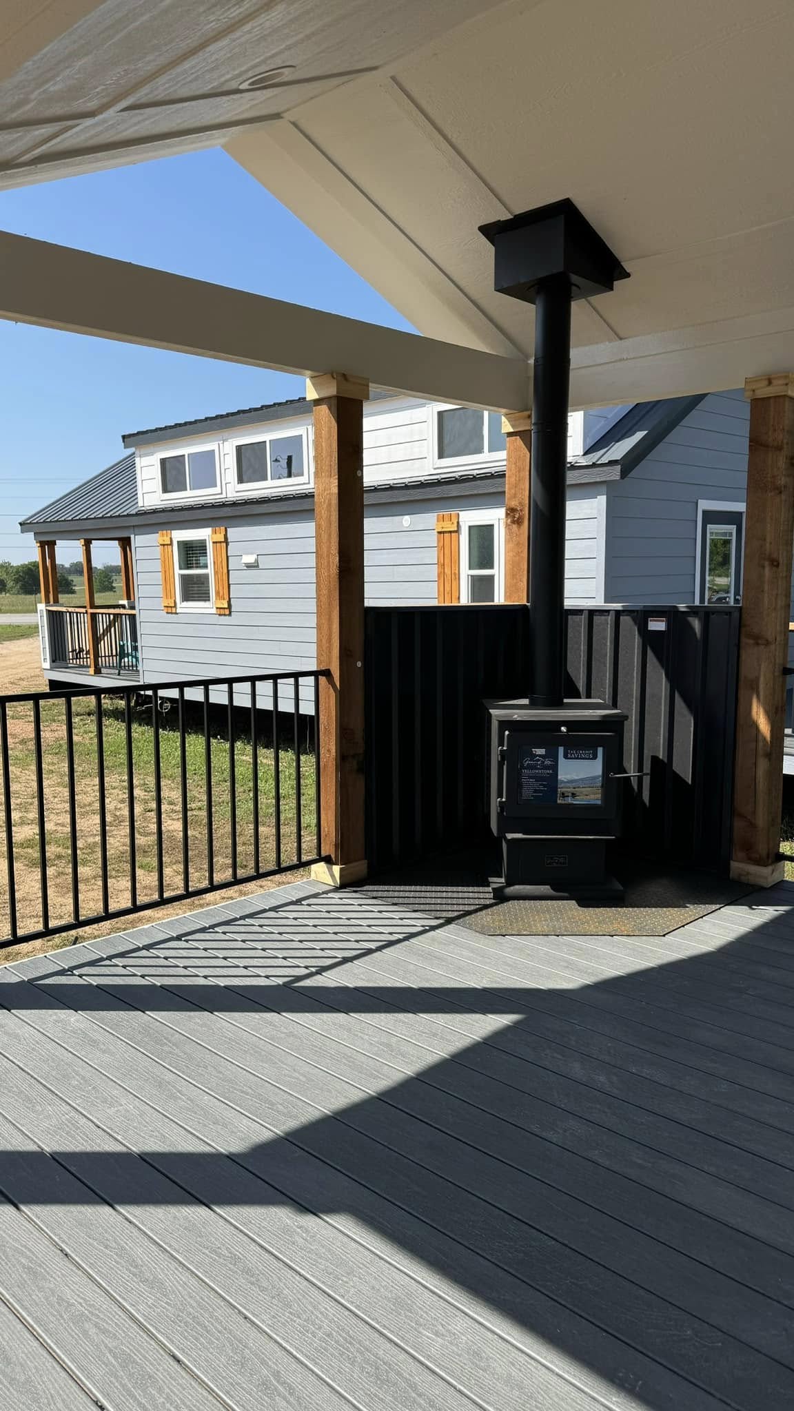 Alt text: A cozy porch with wood beams and a small black stove overlooks a gray tiny house with orange shutters. Sunlight casts shadows on the deck.