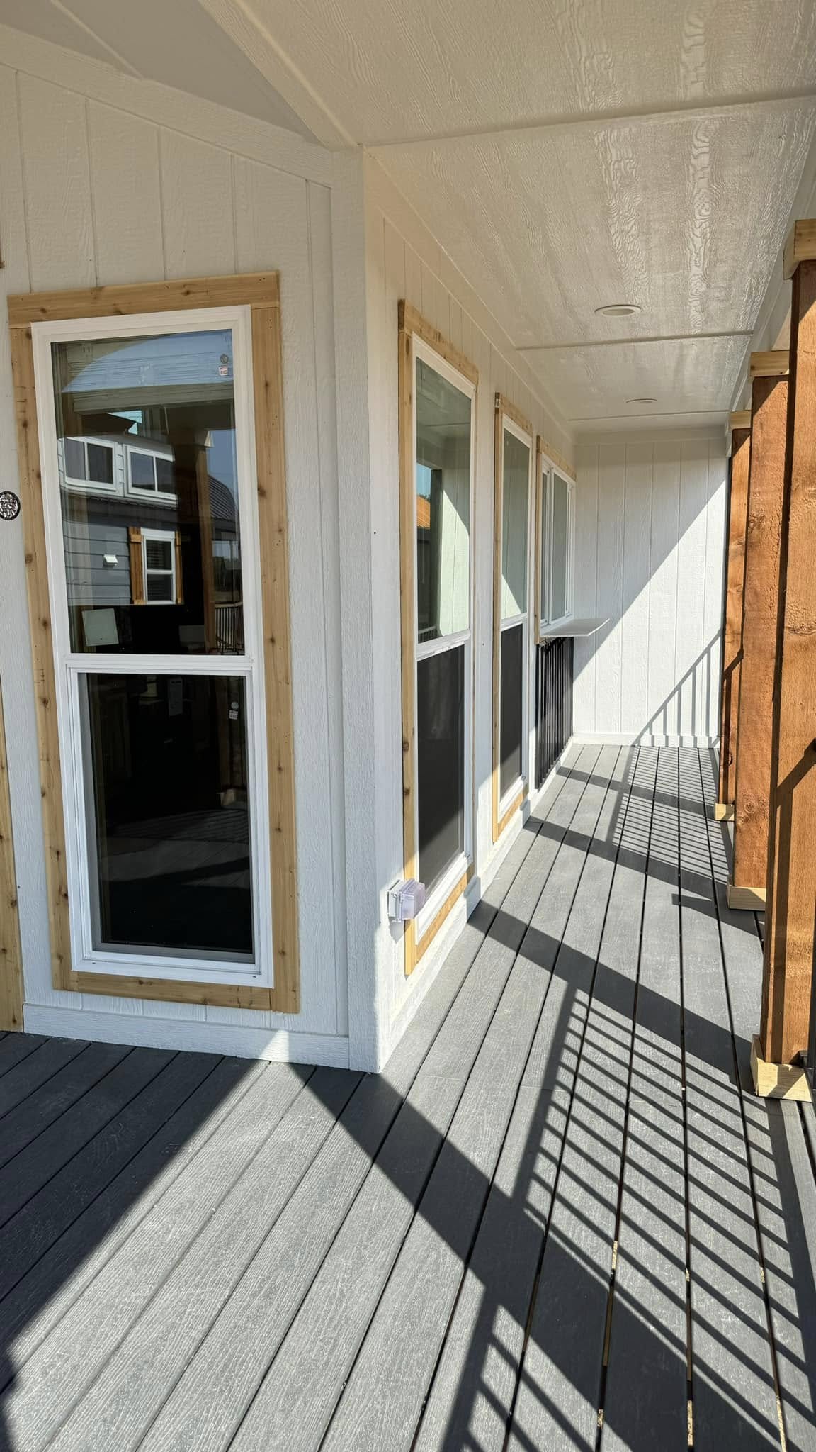 A narrow, sunlit porch with light wood siding and four vertical windows. The wooden railing casts shadows on the gray floor, creating a serene ambiance.