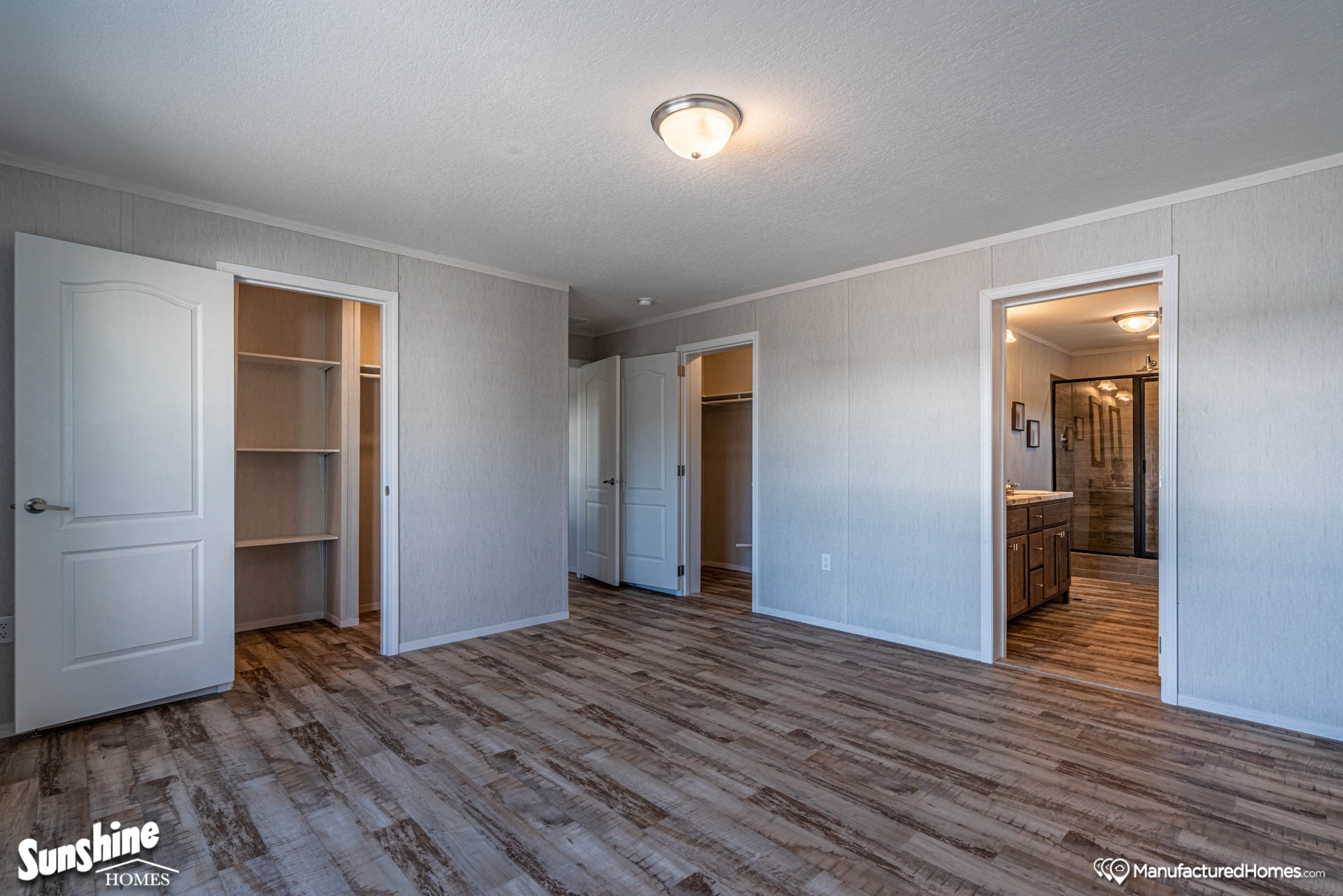 Spacious room with wood flooring, light gray walls, and a ceiling light. Open doors reveal a closet with shelving and a bathroom with a modern design.