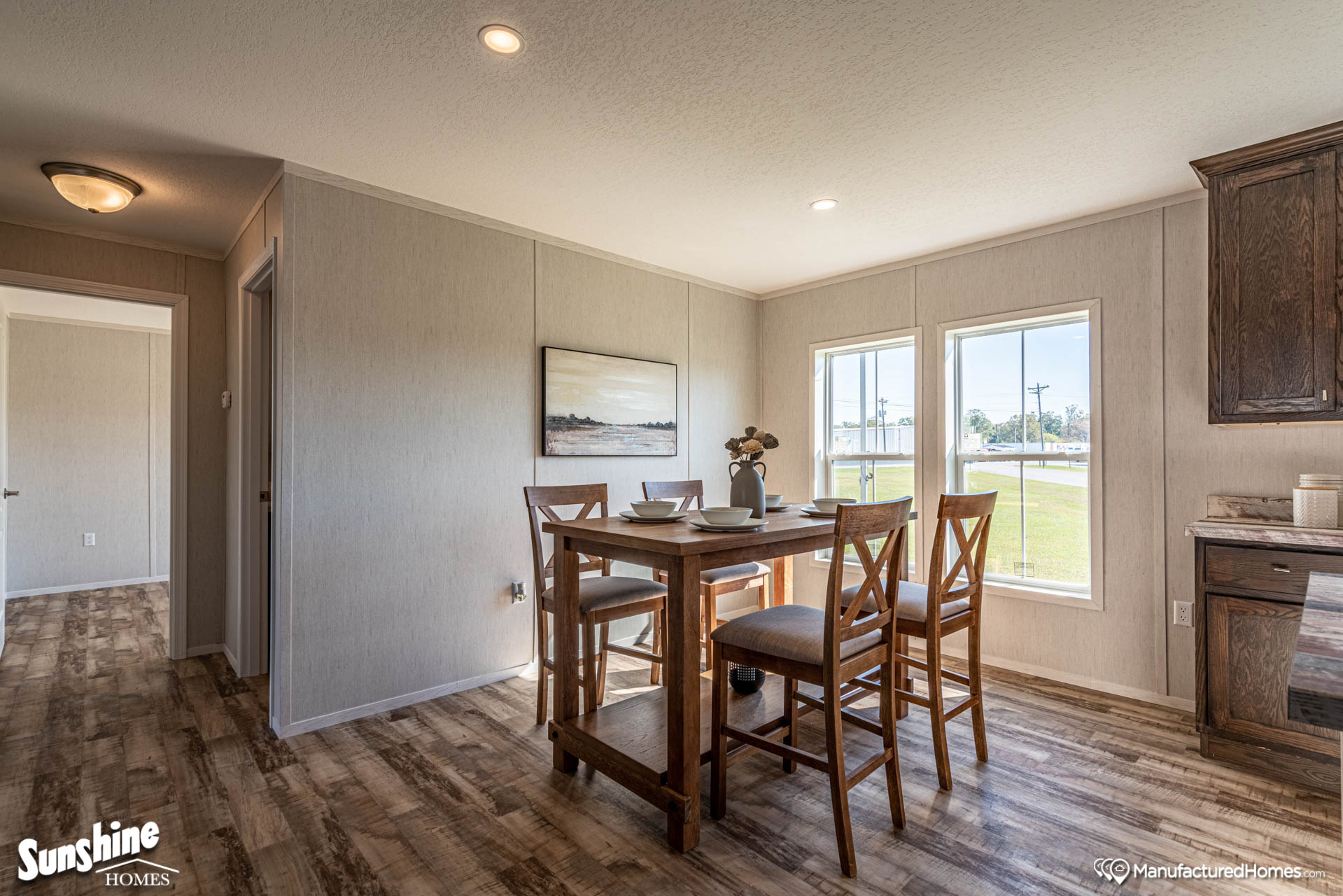 Bright dining area in a manufactured home, featuring a wooden table set with place settings for four, wood-patterned floor, large windows, and neutral wall art. Warm, inviting atmosphere.