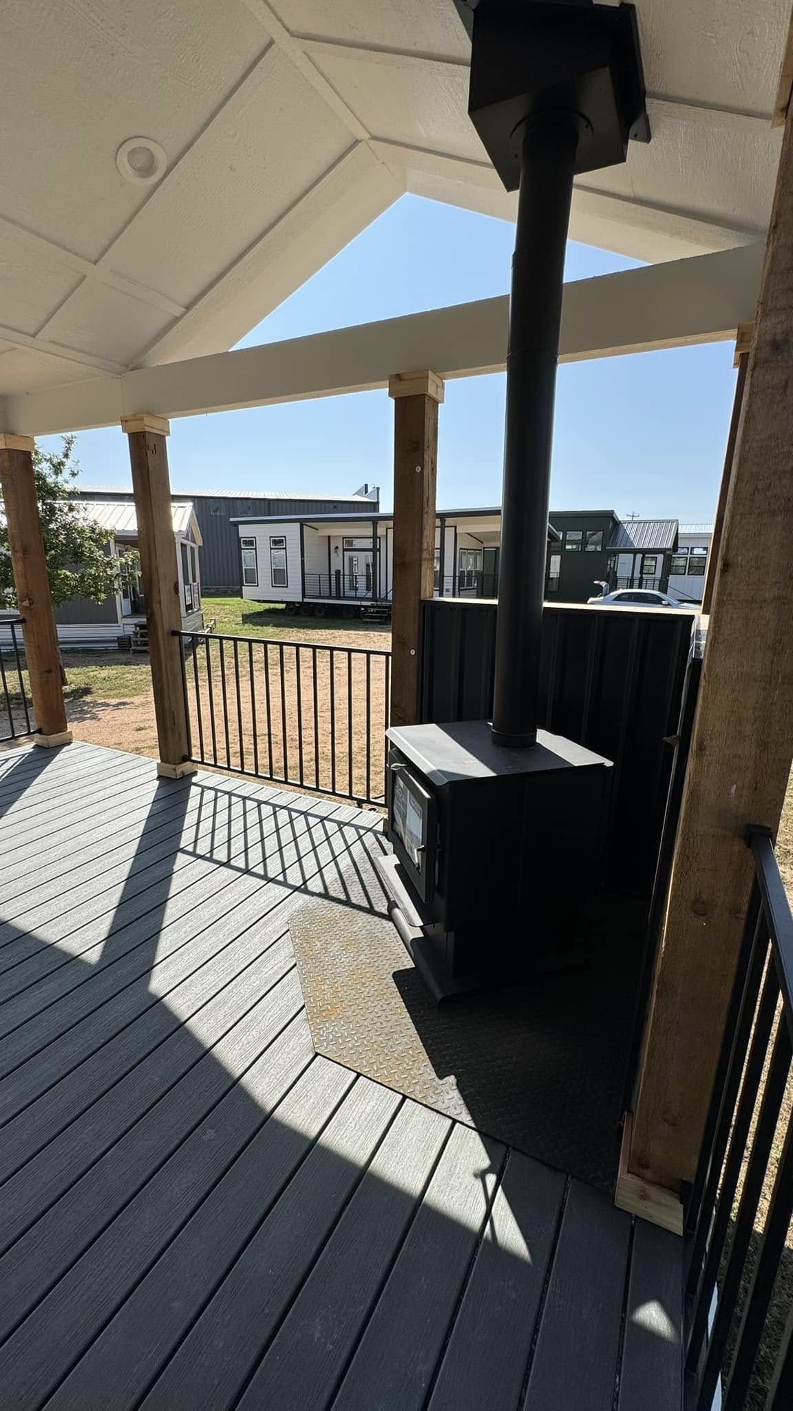 A porch with wooden beams and a slanted roof features a black iron stove and chimney. Sunlight casts geometric shadows on the gray deck. Small houses are visible in the background.
