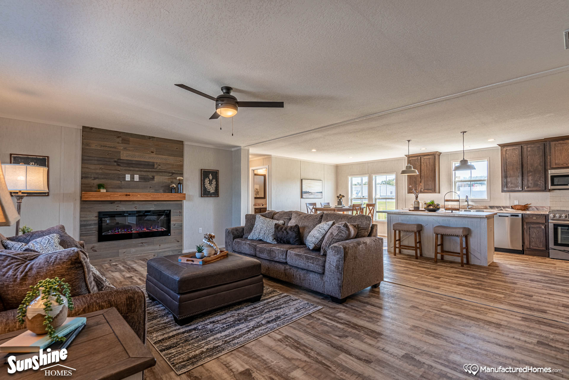 Open living space features a cozy sitting area with a gray sofa, fireplace, and ceiling fan. The adjacent kitchen has wooden cabinets and an island with stools.
