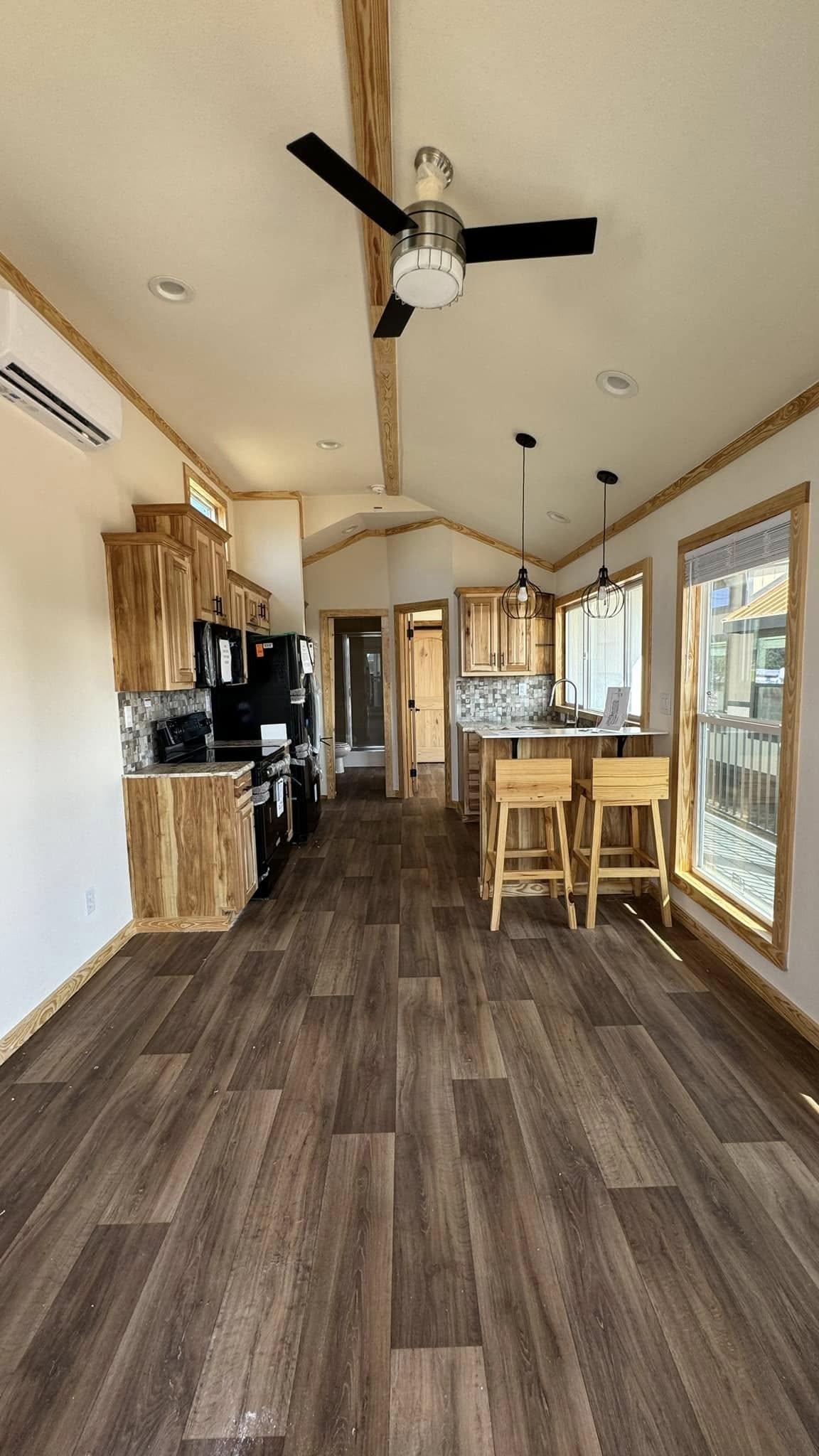 Spacious kitchen with wood cabinets and floors, black appliances, bar stools, and modern light fixtures. Bright natural light from a large window.