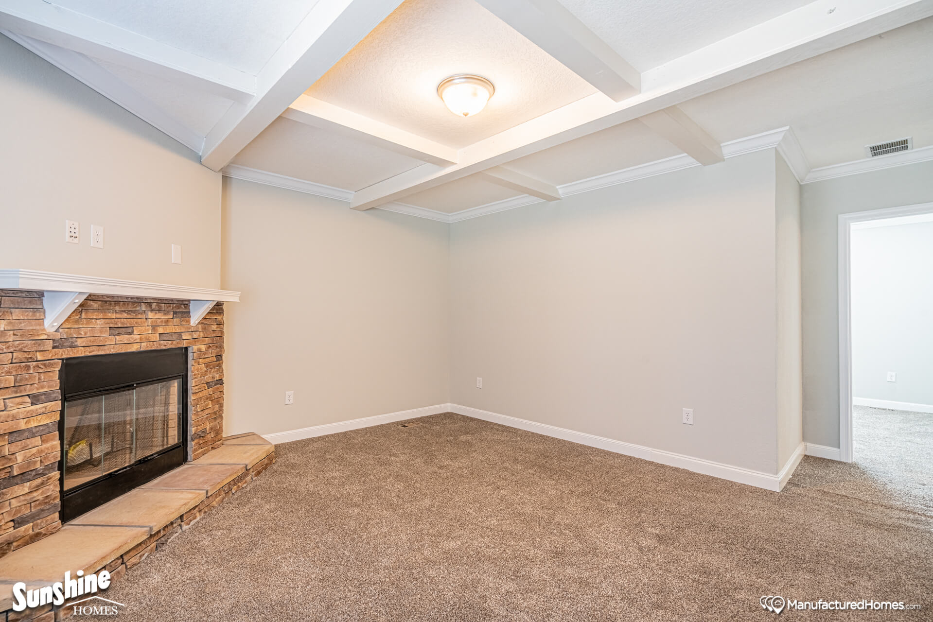 A cozy, empty living room with beige carpet, white walls, and ceiling beams. Features a stone fireplace on the left wall and a ceiling light.