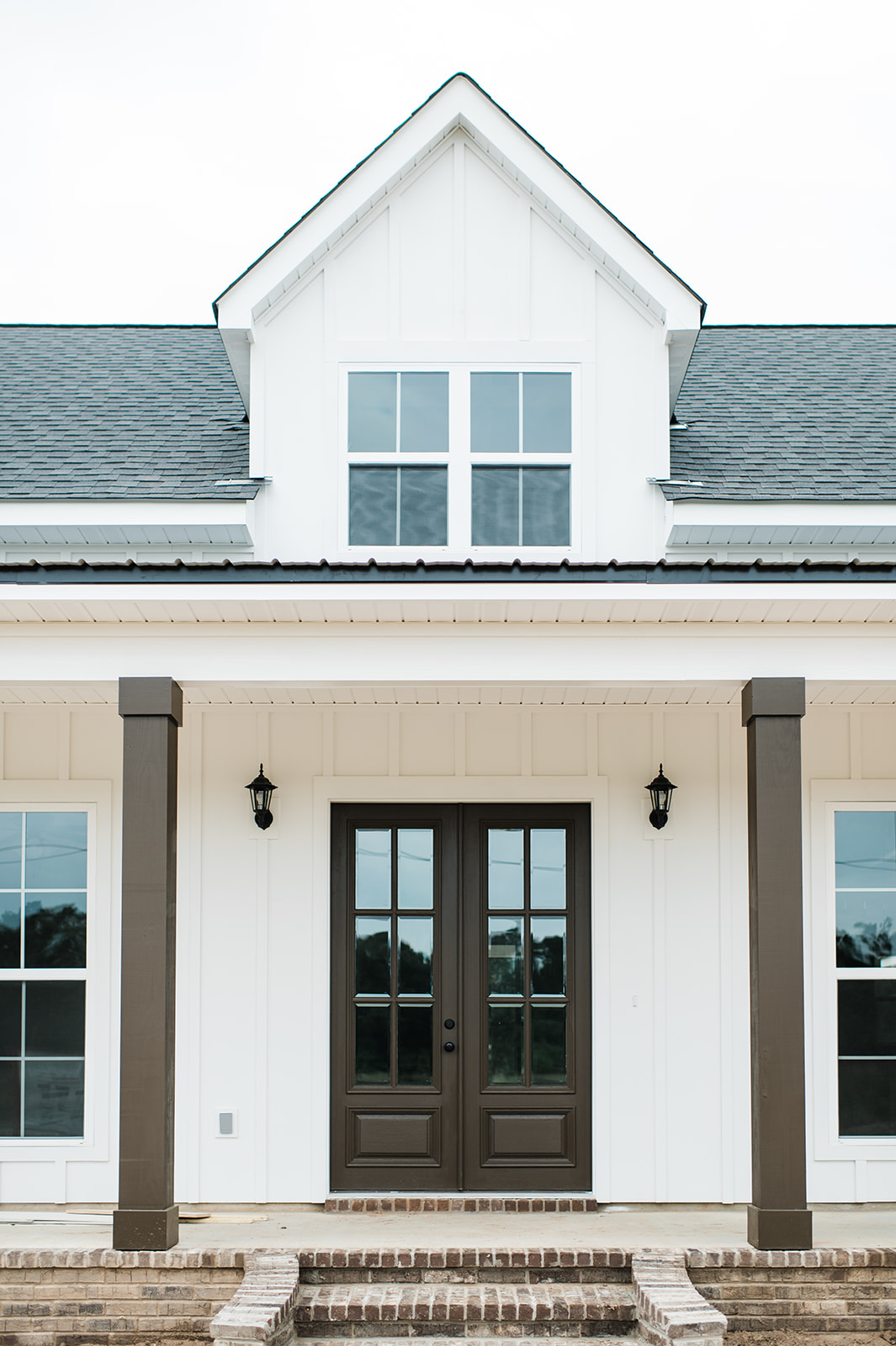 Front view of a modern farmhouse with white siding, dark brown double doors, and twin exterior lanterns. A brick porch and steps lead up to the entrance.