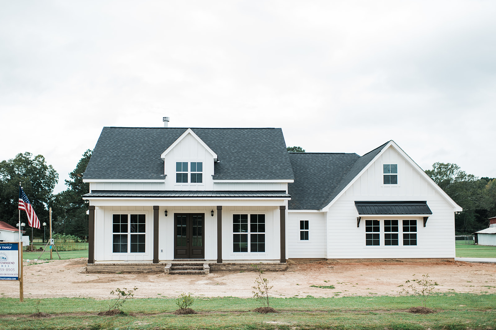 A new, white, farmhouse-style house with black roof shingles stands under a cloudy sky. An American flag and greenery add a welcoming feel.