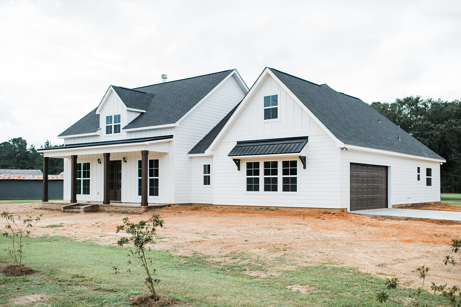 Modern white farmhouse with black roof, large windows, and a spacious porch. The house stands on a sparse lawn, exuding a calm, welcoming vibe.