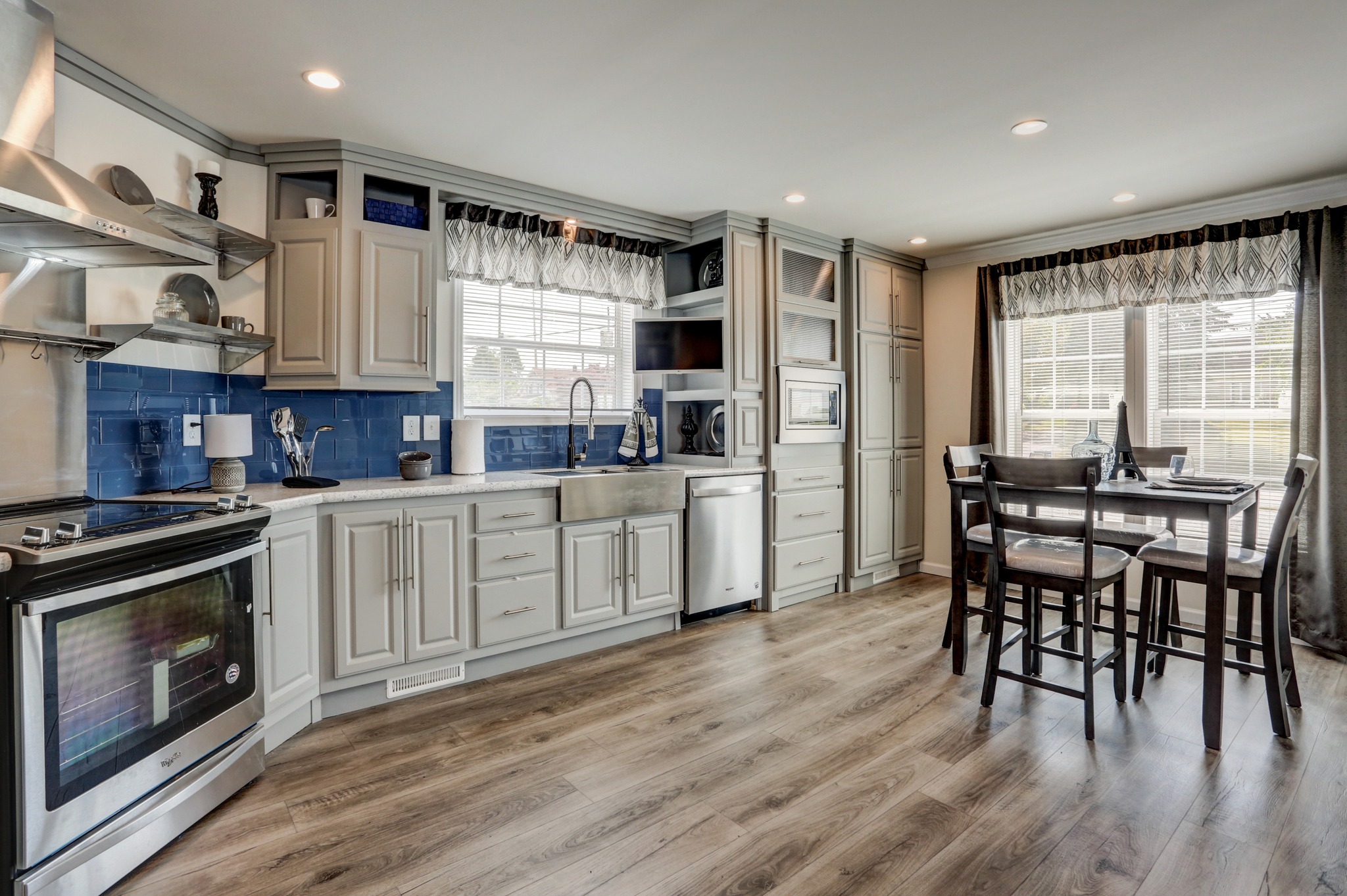 Modern kitchen with gray cabinets, blue backsplash, and stainless steel appliances. A wooden dining table is by large windows, creating a cozy feel.