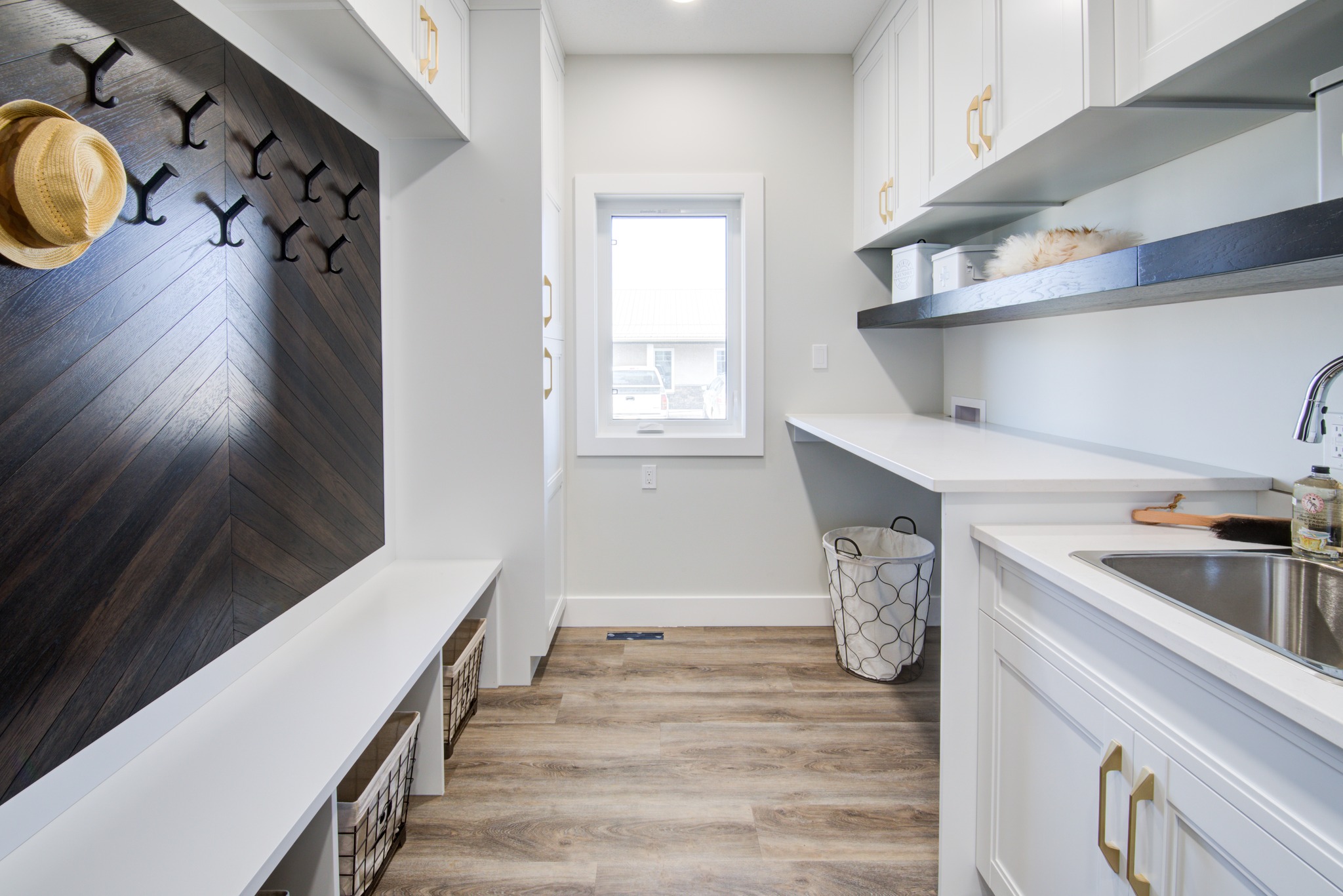 A modern mudroom features white cabinets, a dark wood accent wall with hooks, a bench with baskets, a window, and a sink. The room feels clean and organized.