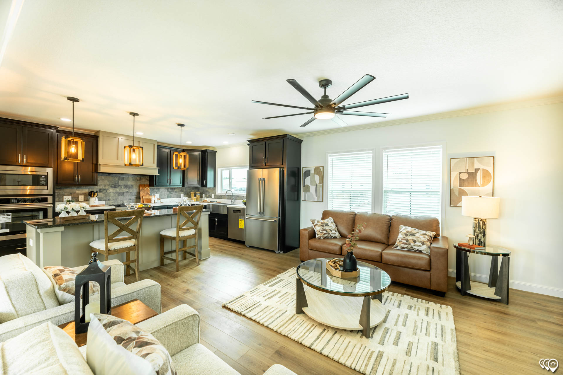 Modern open-concept living room and kitchen featuring light wood flooring, a brown leather sofa, two patterned chairs, and a stylish kitchen island with pendant lights.