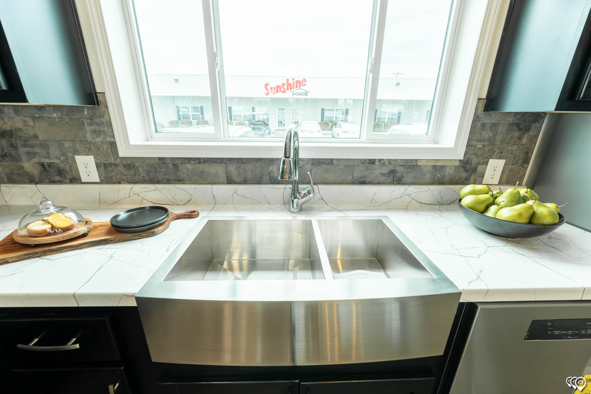 A modern kitchen sink with dual basins is set in a marble countertop. On the left, a wooden board holds pastries under a glass dome. On the right, a bowl of green pears adds a fresh touch. A large window behind the sink offers a view of a building with the "Sunshine" sign visible.
