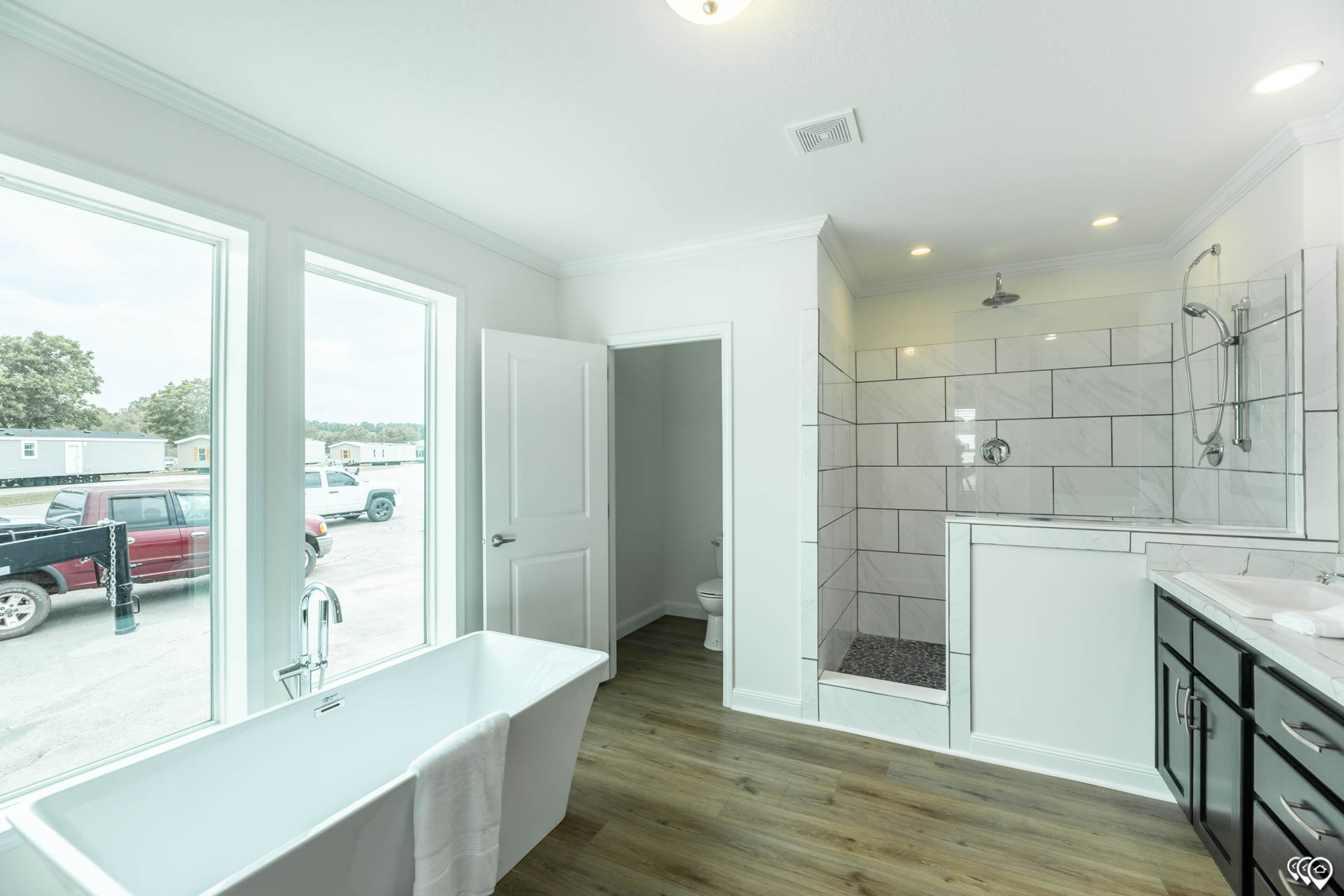 Spacious bathroom with a freestanding tub near large windows, a walk-in shower with white tiles, dark wood cabinets, and wood flooring. Bright and modern ambiance.