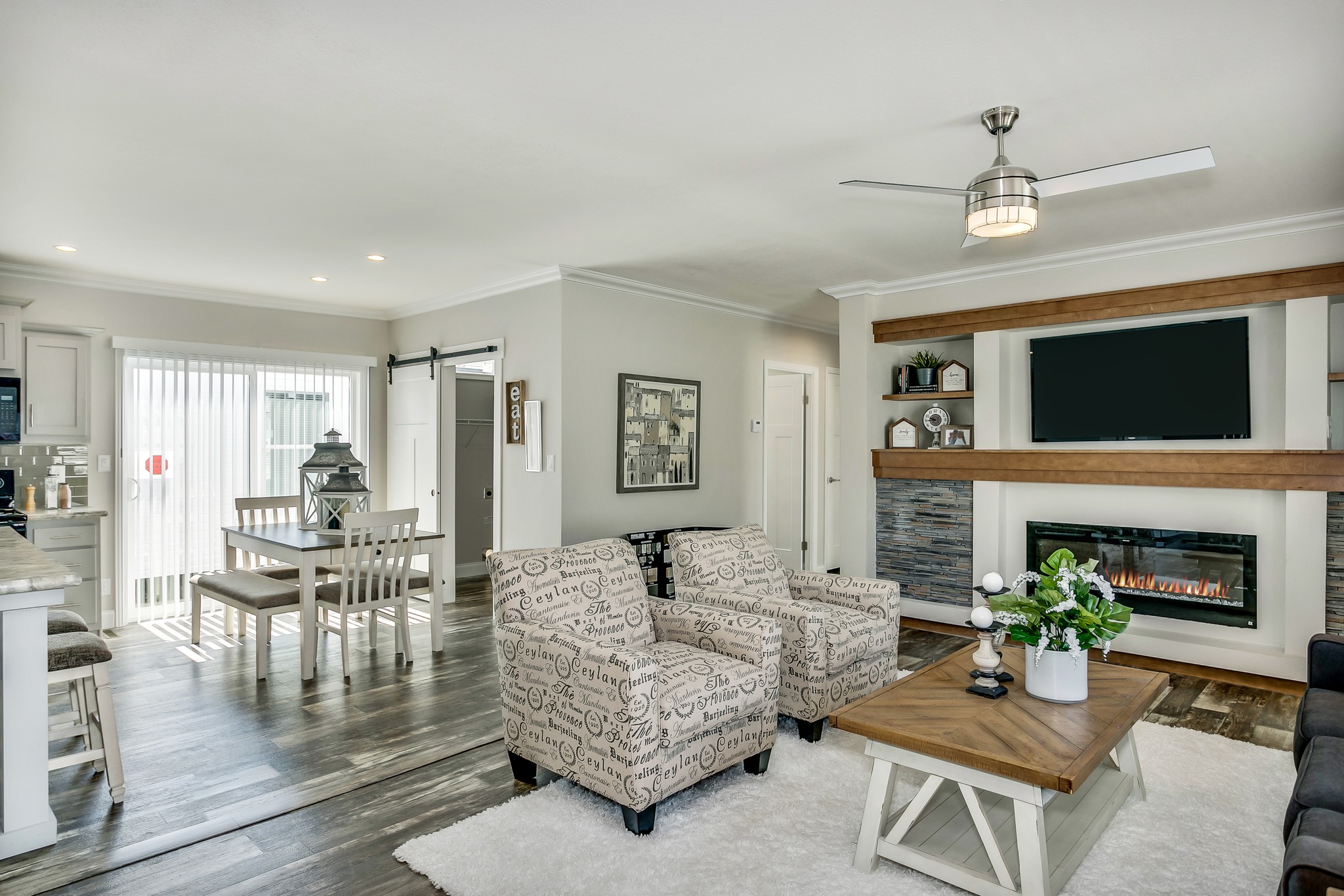 Modern living room with light neutral tones. Features two patterned armchairs, a wooden coffee table, a flat-screen TV above an electric fireplace, and an open dining area with sliding glass doors. Bright and welcoming atmosphere.