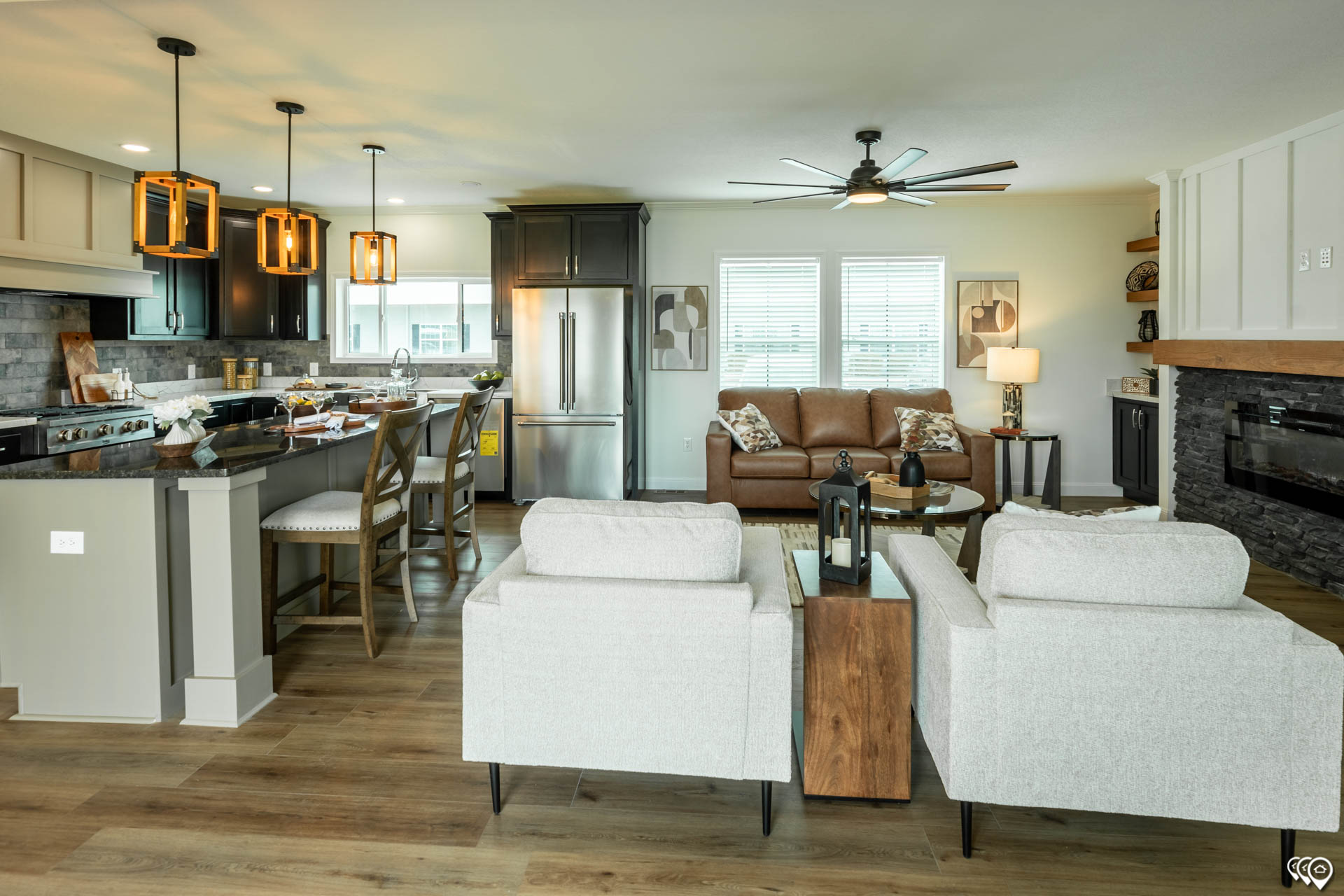 Modern open-concept living room and kitchen with wooden floors. Features a cozy brown sofa, light armchairs, pendant lights, and sleek kitchen appliances.