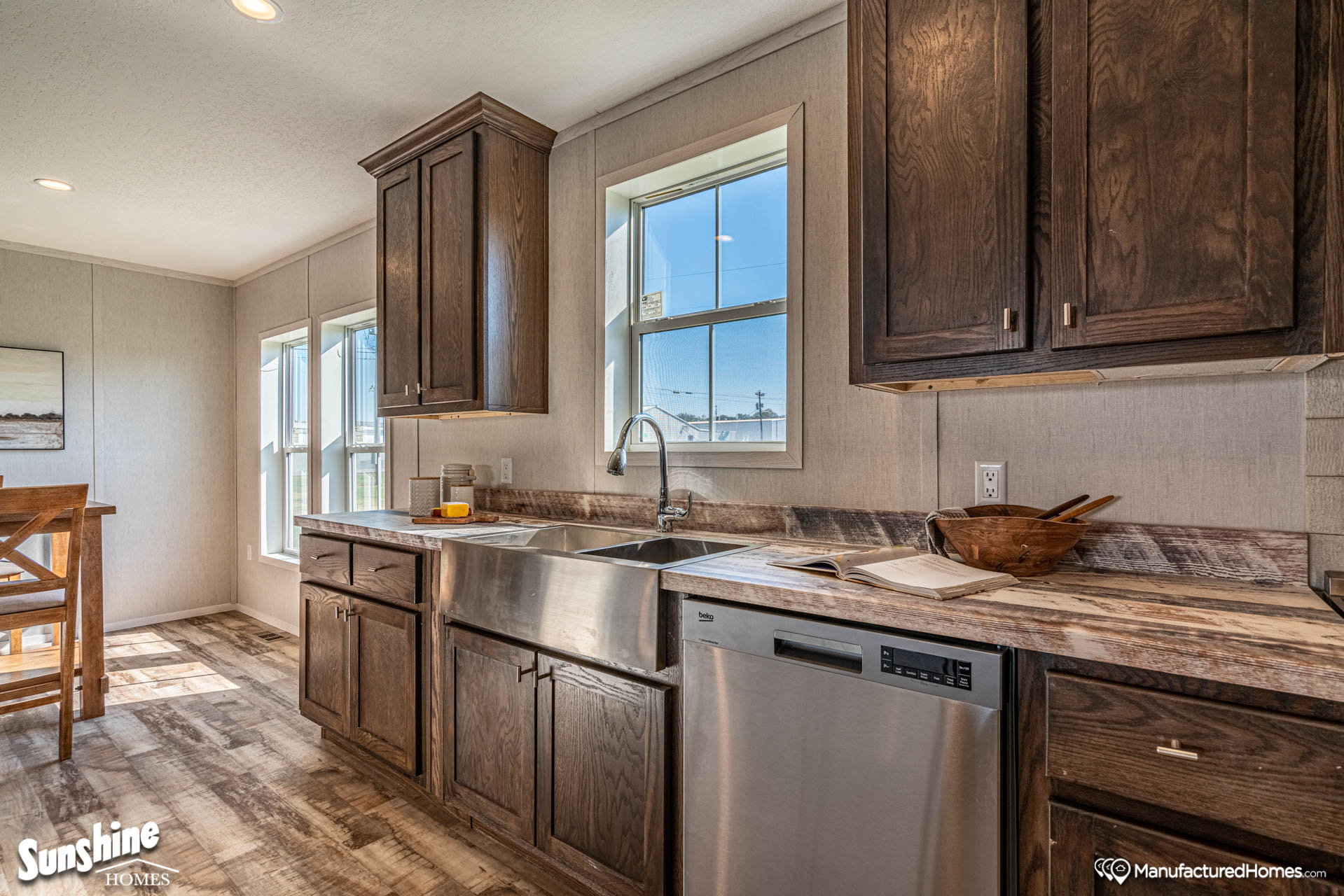 Bright kitchen with dark wooden cabinets, stainless steel farmhouse sink, and dishwasher. Sunlight streams through windows, creating a warm ambiance.