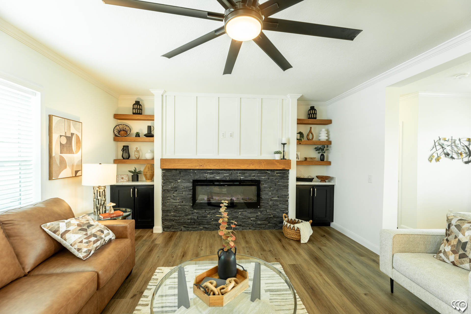 Modern living room with a beige couch, geometric pillows, and a glass table. Features a stone fireplace, wooden shelves, and a ceiling fan. Cozy and elegant.