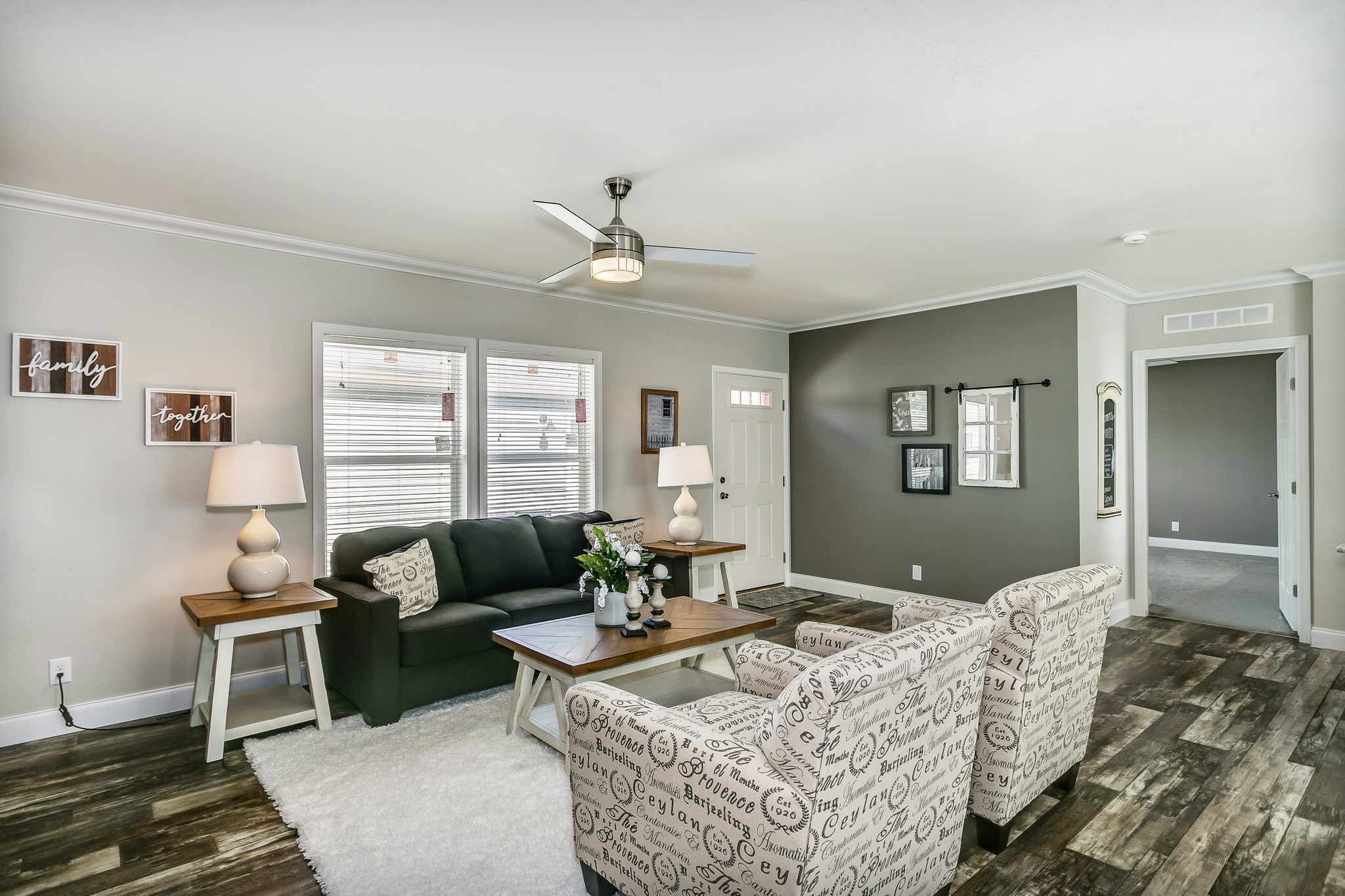 A cozy living room with a dark green sofa, two patterned armchairs, and a wooden coffee table. Lamps and family-themed wall art add warmth. Neutral tones.