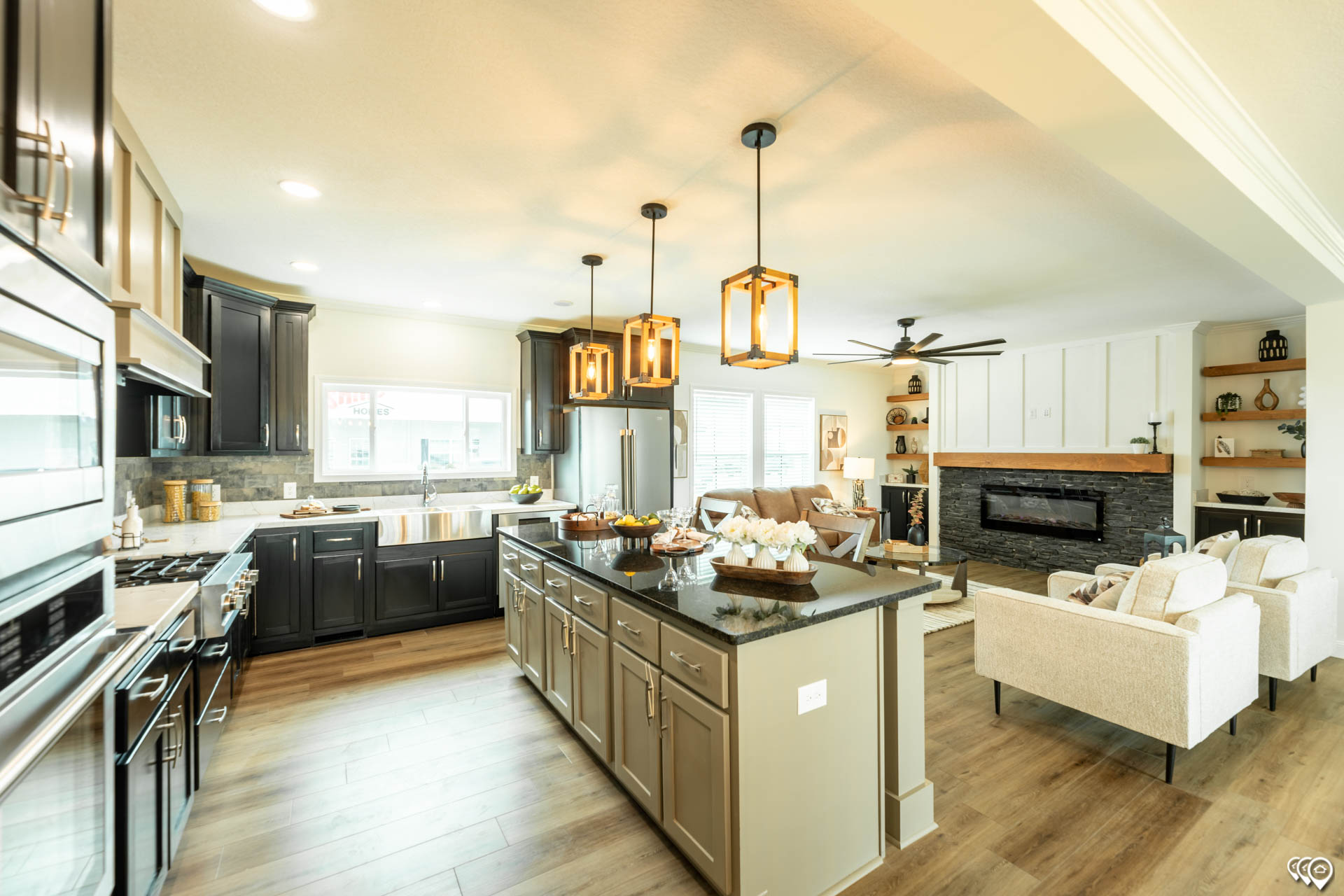 Modern kitchen and living area with open layout. Features an island with pendant lights, dark cabinets, white sofas, and a stone fireplace, conveying warmth.