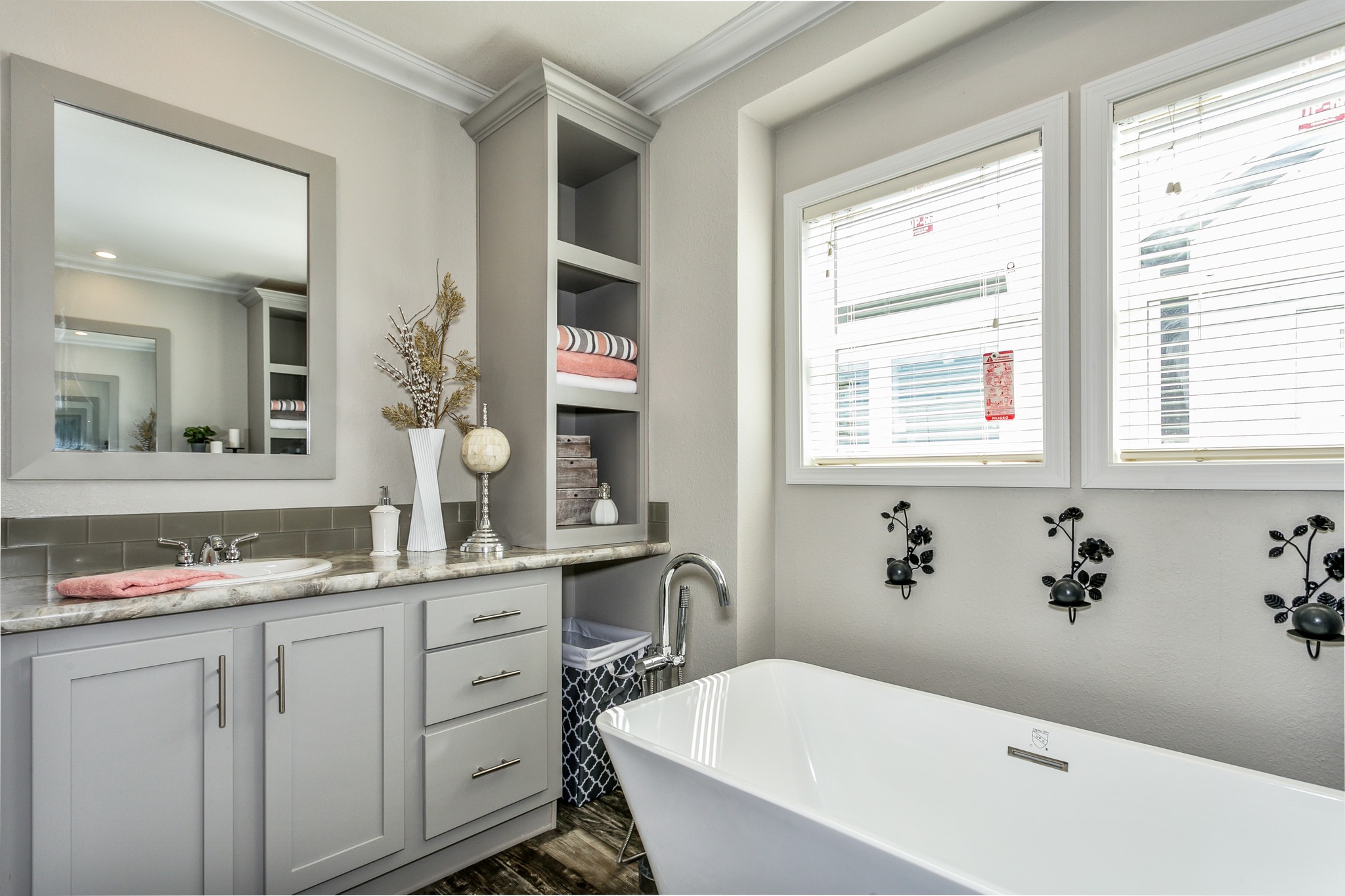 Modern bathroom with white freestanding tub, gray vanity, large mirror, open shelves with towels, decorative plants, and light streaming through blinds.