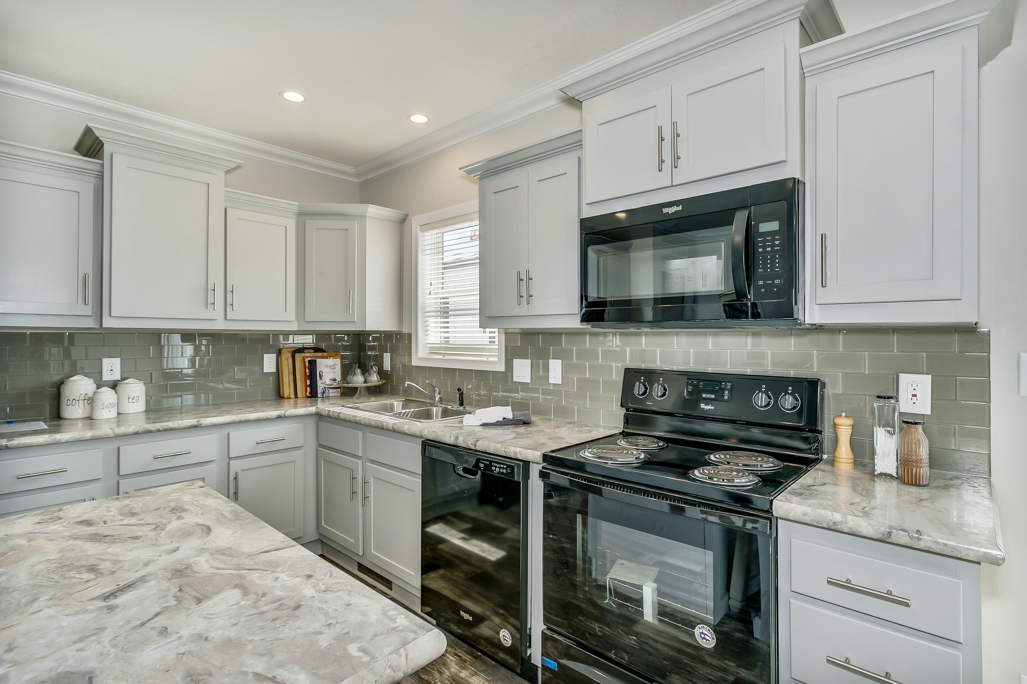 Modern kitchen with light gray cabinets, marble countertops, and stainless appliances. A window brings in natural light, enhancing the clean, fresh ambiance.