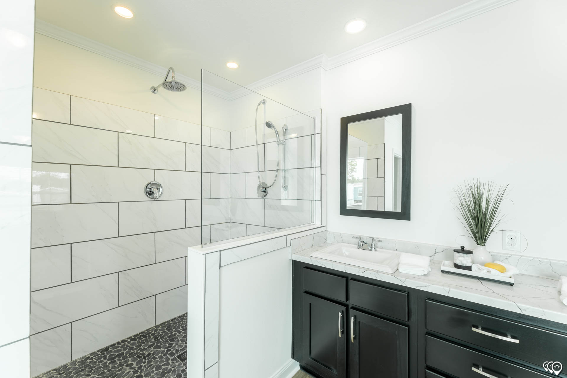 Modern bathroom with a glass-enclosed shower. White subway tiles line the shower, with a stone floor. The sink has a dark vanity, mirror, and decor.