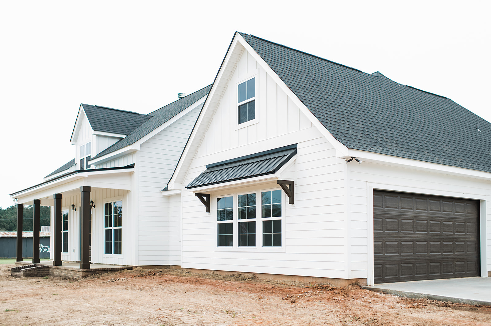 A modern white farmhouse with a dark roof, multiple windows, and a two-car garage. The front porch is supported by dark wooden pillars, conveying a classic, serene ambiance.