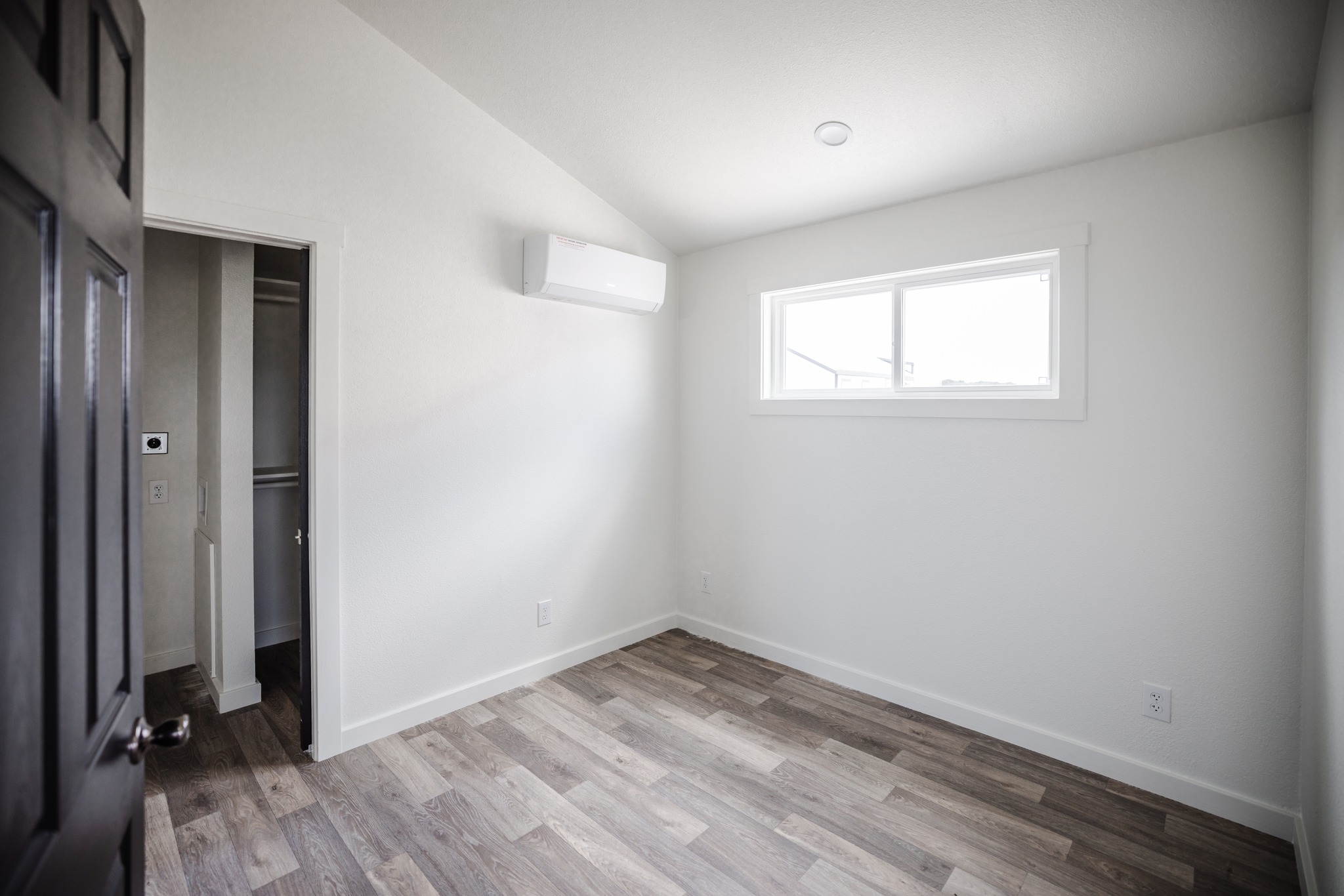 Minimalist empty room with white walls and wooden floor, featuring a small window on the right, an air conditioner above, and an open closet door on the left.