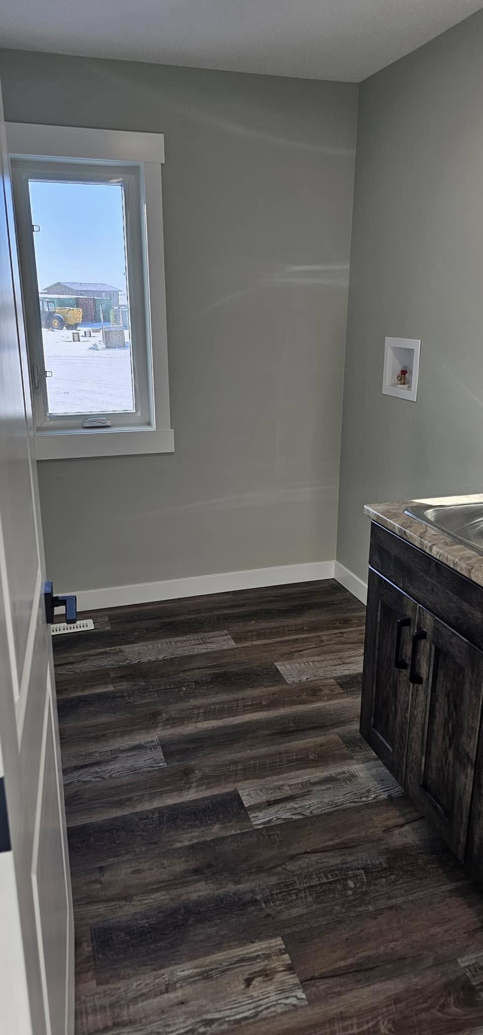 Small utility room with light gray walls and a window showing an outdoor landscape. Dark wooden flooring complements a sink with a wooden cabinet.