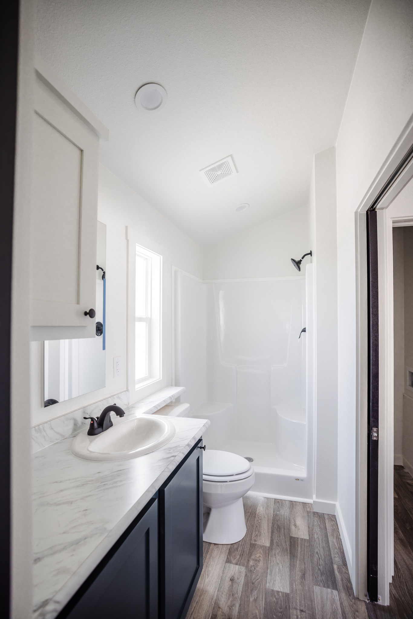 Narrow modern bathroom with white walls, wood floor, and a marble countertop. Includes a white sink, black cabinet, toilet, shower, and natural light from a window.