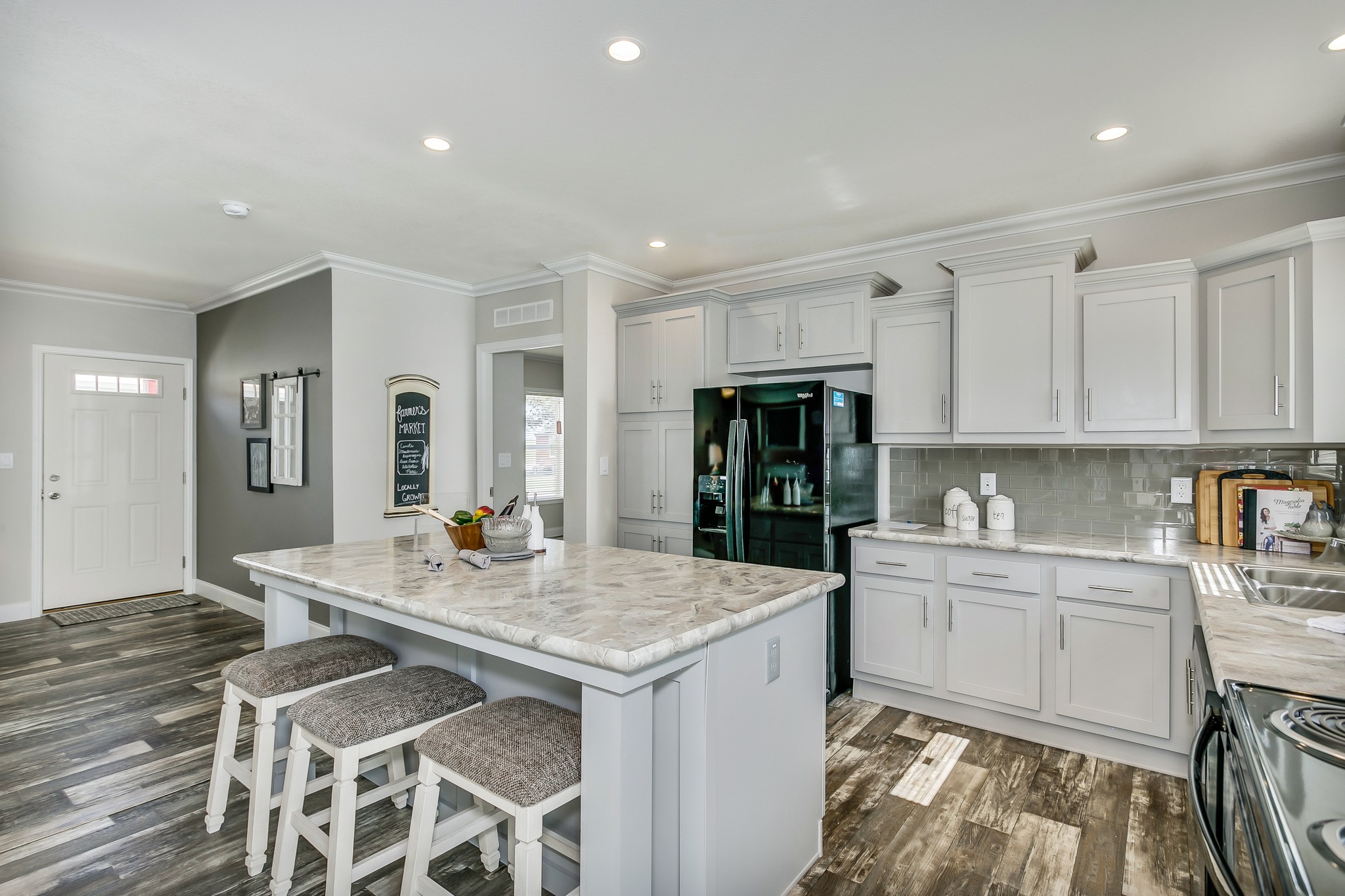 Modern kitchen with granite island, three cushioned stools, white cabinets, stainless steel appliances, and grey wood flooring. Bright, inviting ambiance.