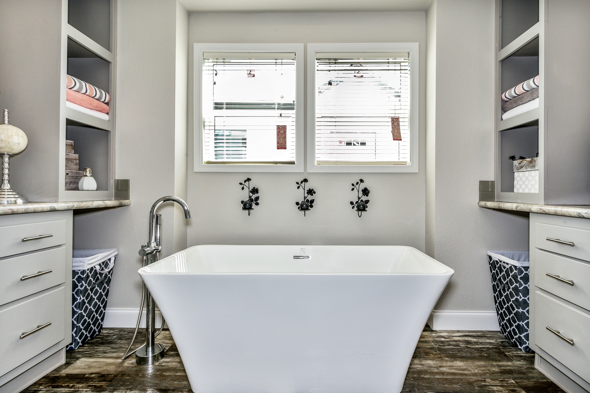 Luxurious bathroom with a white freestanding tub beneath two windows. Shelves with towels and decorative items on both sides. Elegant and serene atmosphere.