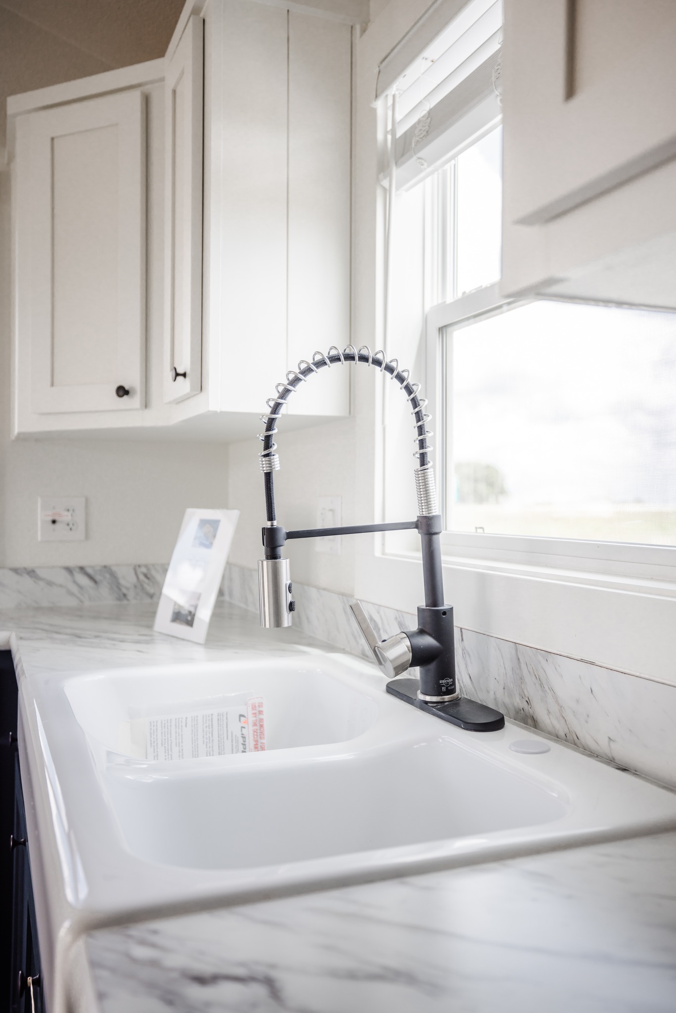 Modern kitchen sink with a sleek, high-arc faucet and marble countertop. White cabinets and sunlight streaming through a nearby window create a fresh, bright atmosphere.