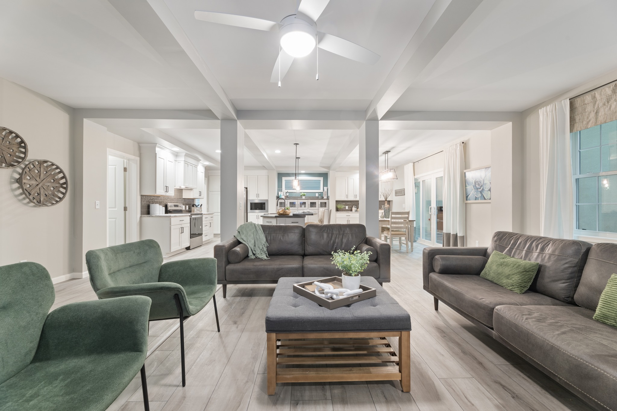 Modern living room with gray sofas and green chairs around a wooden coffee table. A ceiling fan, neutral tones, and open kitchen create a cozy, airy feel.
