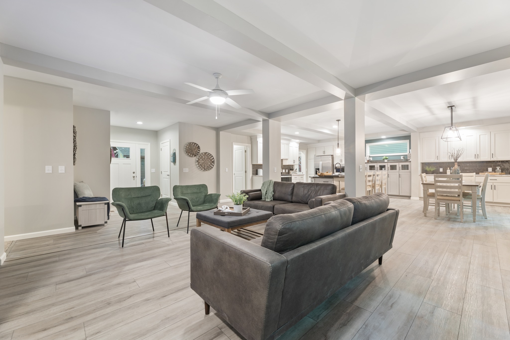 Modern open-concept living space with gray sofas, green chairs, and a wooden coffee table. Light wood flooring, kitchen area with white cabinets, and dining set. Cozy and welcoming.