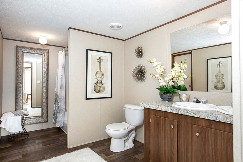A modern bathroom featuring a vanity with a granite countertop, a large mirror, and an orchid plant. There’s a stylish wall art of a violin, a toilet, and decorative accents on the walls. The wooden floor and natural lighting create a warm, serene atmosphere.