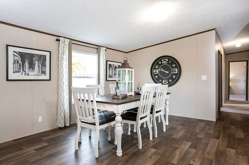 Bright dining room with a wooden table and six white chairs on a hardwood floor. A large black wall clock, framed photos, and a lantern centerpiece add elegance.