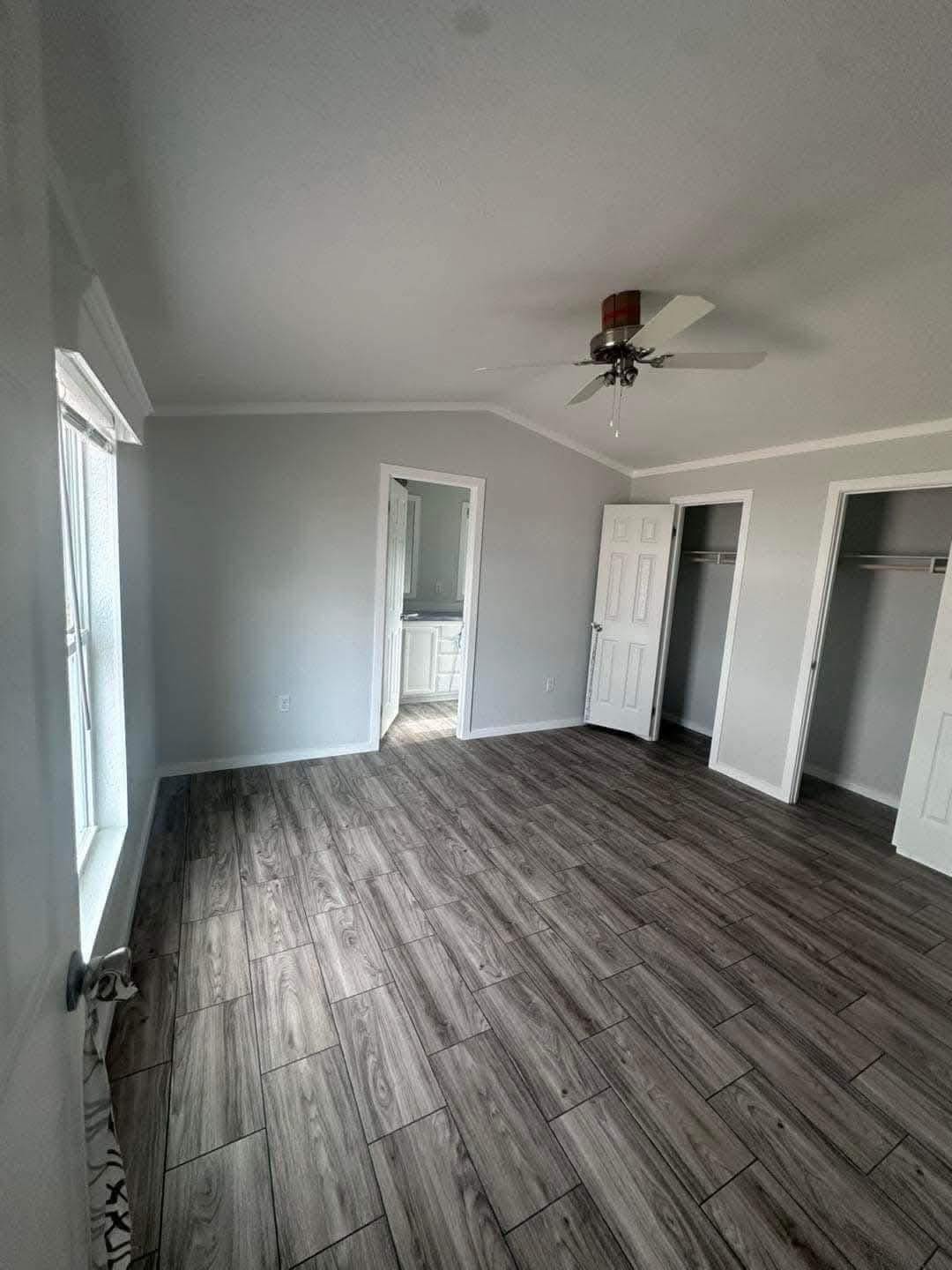 Empty room with gray walls and wood-patterned floor tiles. Two open closets and a door leading to a bathroom. Ceiling fan hangs in the center. Bright and minimalistic.