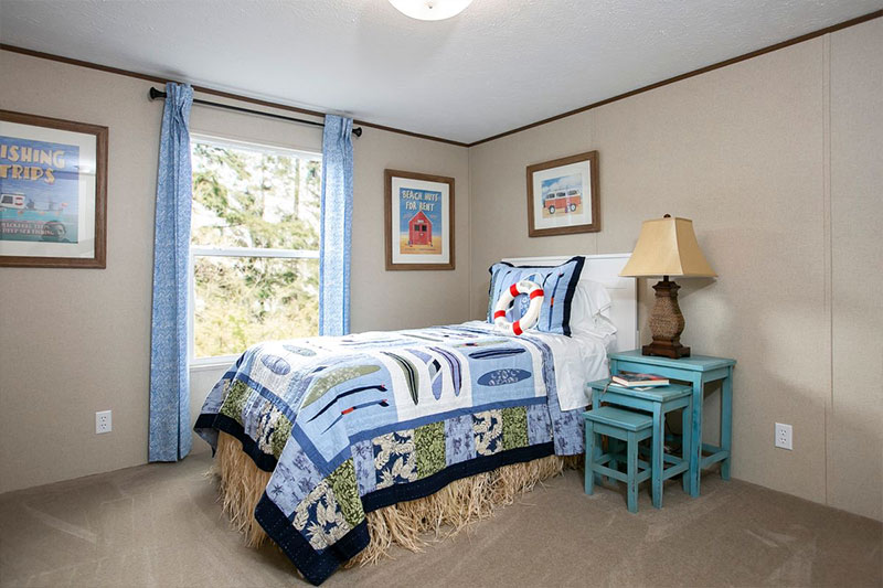 A cozy bedroom with a nautical theme features a bed with blue patterned bedding and a life preserver pillow. Light blue curtains frame a window, and two framed seaside prints hang on beige walls. A turquoise bedside table with a lamp completes the peaceful ambiance.