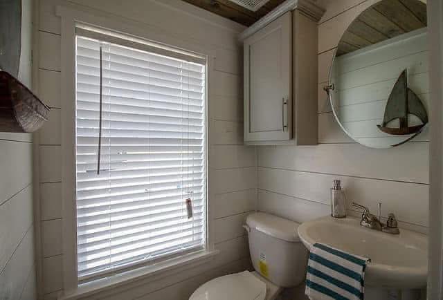 Compact bathroom with white shiplap walls, circular mirror, striped towel, and boat model. Sunlight filters through white blinds, creating a cozy feel.