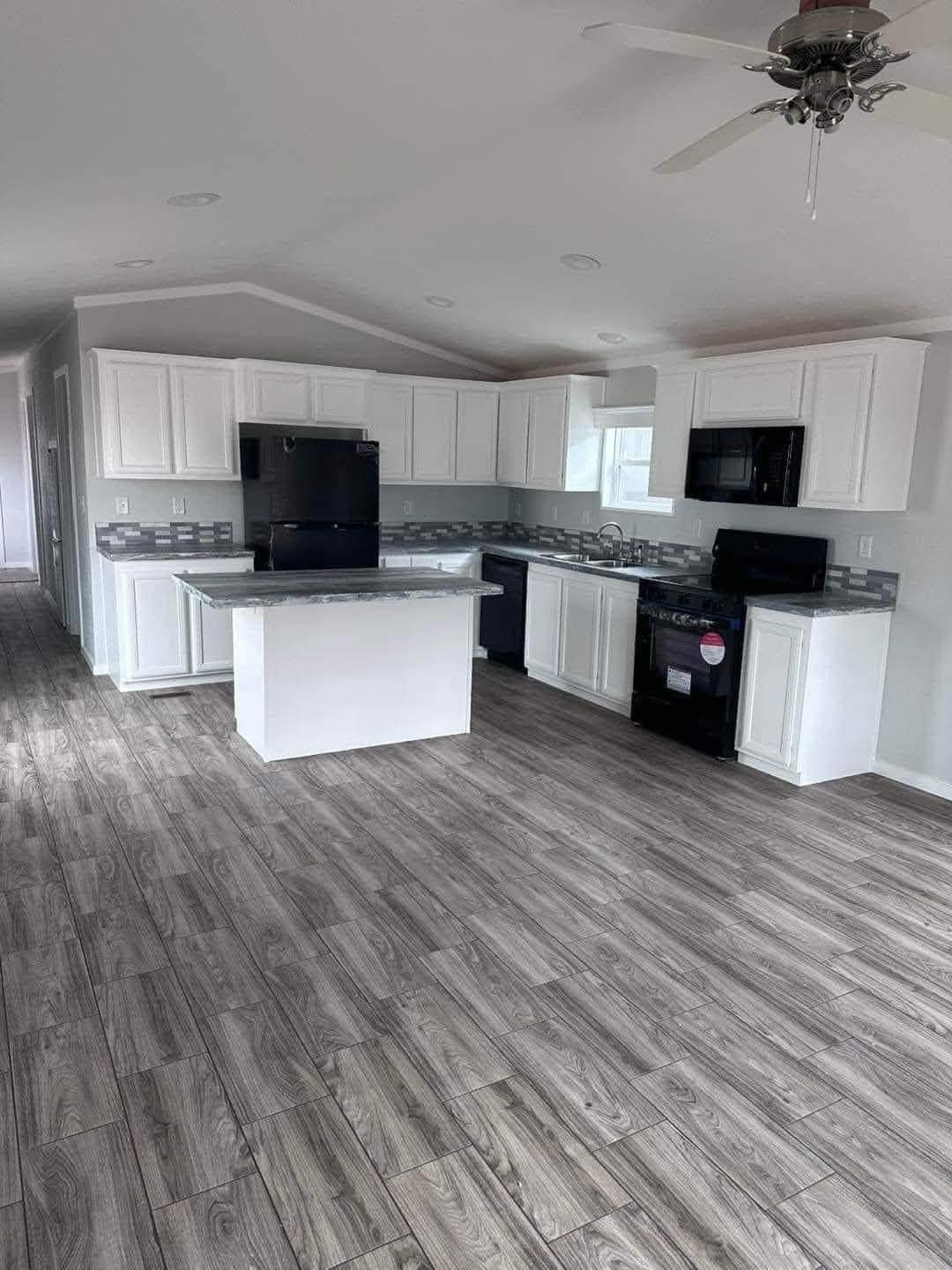 Spacious kitchen with white cabinets, dark appliances, and gray wood-like flooring. Central island with marble countertop. Bright and modern feel.