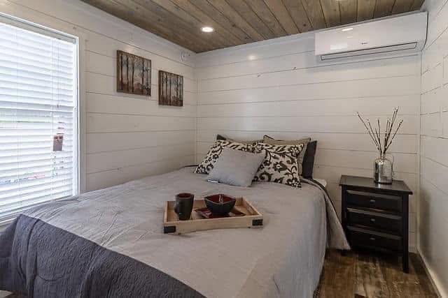 Cozy bedroom with a wooden ceiling and white shiplap walls. Features a neatly made bed with patterned pillows, a side table, and nature-themed decor.