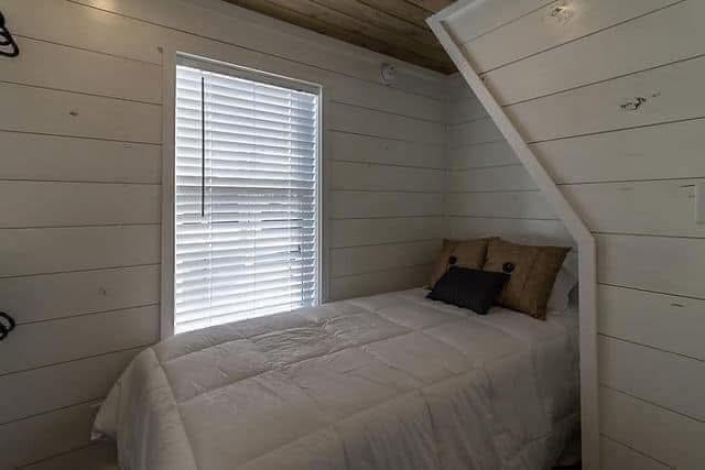 Cozy bedroom with white shiplap walls, featuring a single bed with a white comforter and accent pillows. A window with blinds lets in soft light.