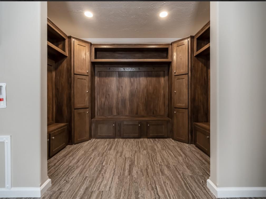 A spacious mudroom with rich wooden cabinetry and hooks for hanging items. Overhead lighting brightens the wood grain floor and the room's warm tones.