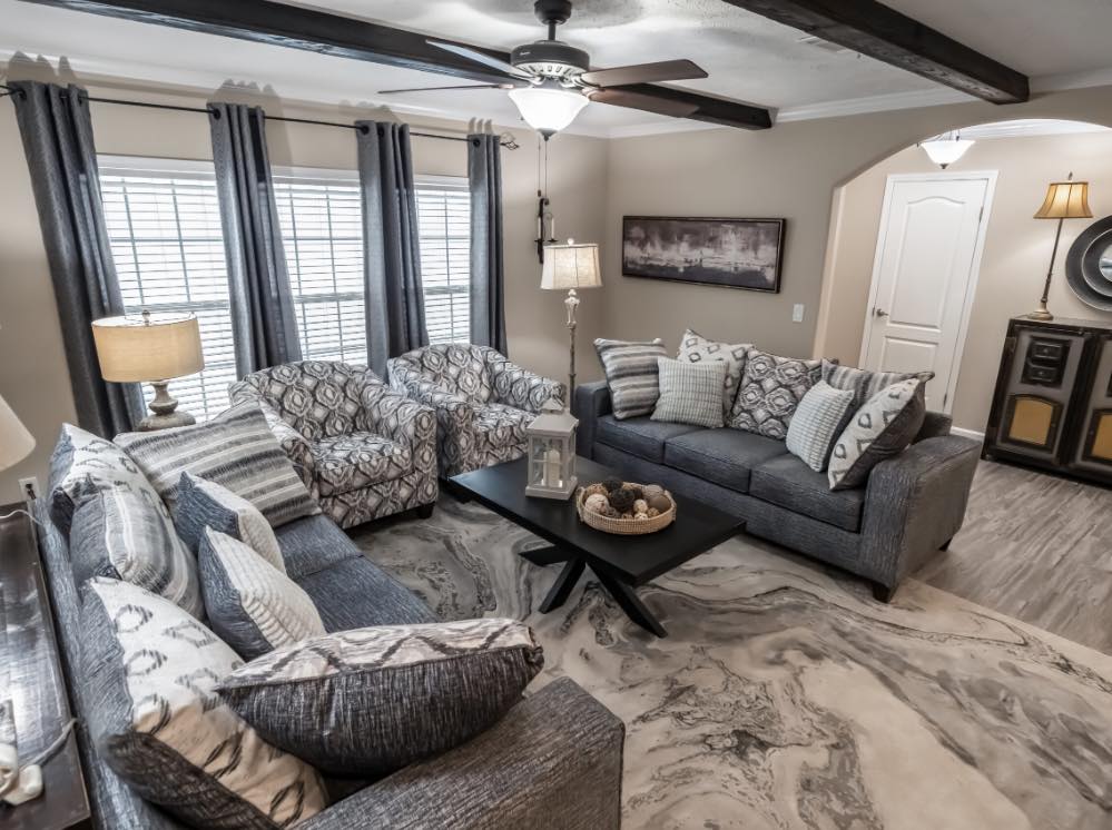 Cozy living room with a modern design features a gray sectional sofa, patterned armchairs, and a marble rug. Natural light filters through large windows.