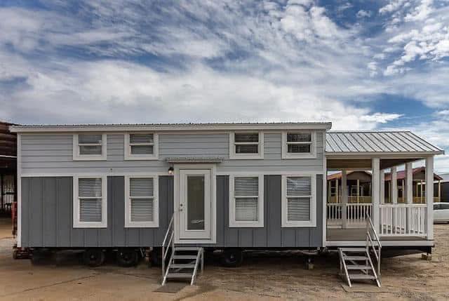 Gray and white tiny house on wheels under a partly cloudy sky. Features a small porch with railings and steps, creating a cozy and inviting atmosphere.