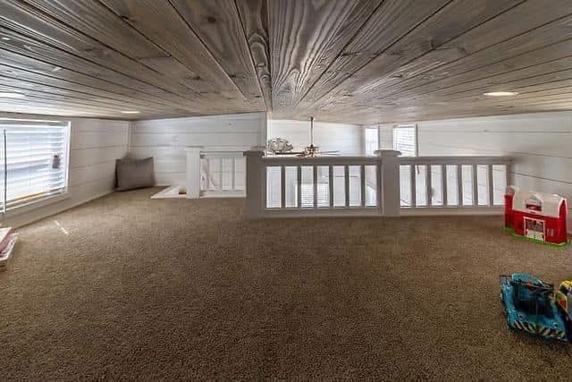 Low-ceiling attic playroom with white walls, brown carpet, and wooden ceiling. Toys are scattered, and a small window offers soft daylight. Cozy and inviting.