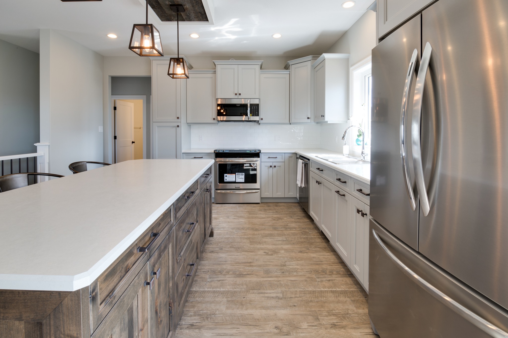 Modern kitchen with light gray cabinets, stainless steel appliances, large island with wood base, pendant lights, and wooden floor. Bright and spacious.