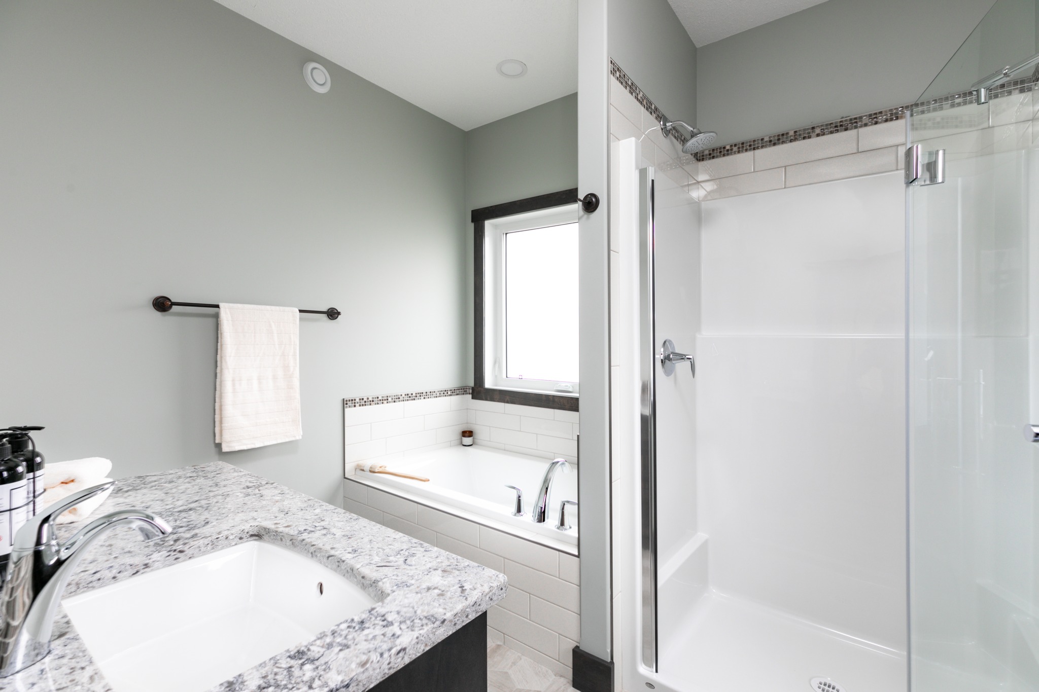 Modern bathroom featuring a granite countertop sink, white bathtub, glass shower, and soft green walls. Bright and clean with natural light.
