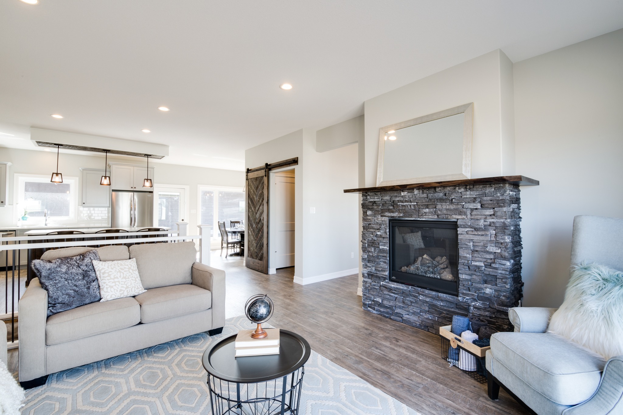 Modern living room with neutral tones features a stone fireplace, beige sofa, and light gray armchair. Open kitchen with pendant lights in background.