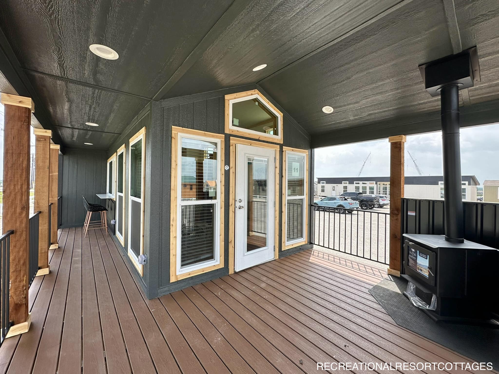 Covered wooden porch with dark siding, large windows, a glass door, and a wood stove. Stools by a bar table overlook parked vehicles and distant buildings.